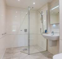 A modern bathroom with a walk-in glass shower, wall-mounted sink, mirror, towel radiator, and beige tiled walls and floor.