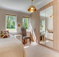 A tidy bedroom with a single bed, a chair, a bedside table with a lamp, large mirrored wardrobes, and two windows letting in natural light.