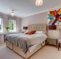 A bright bedroom with a double bed, two side tables, a gray chair, large windows, and colorful abstract art above the headboard.
