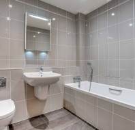 Modern bathroom with gray tiled walls, a wall-mounted sink, a rectangular mirror with lights, a toilet, and a bathtub with a handheld shower.