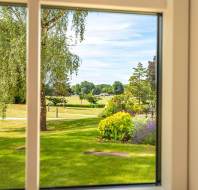 View through a window showing a well-maintained garden with green lawn, flowering shrubs, trees, and a park-like landscape under a blue sky.