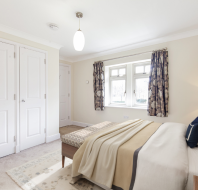 Bright, tidy bedroom with a double bed, beige walls, two windows with patterned curtains, a bedside lamp, and built-in white closets.
