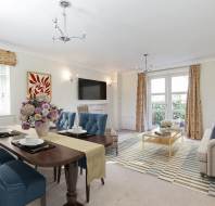 A bright living and dining room features a wooden table with blue chairs, a sofa, a wall-mounted TV, large windows, and floral decor.