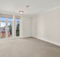 Empty, carpeted room with white walls, a ceiling light fixture, and large windows overlooking a building and trees outside.