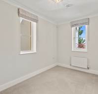 Empty, carpeted room with two windows, cream-colored walls, white trim, a small radiator, and ceiling light fixture. Daylight is visible through the windows.