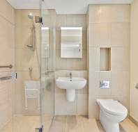 Modern accessible bathroom with beige tiles, wall-mounted sink, toilet, shower area with glass partition, foldable shower seat, grab bars, and a mirrored cabinet.