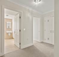 Bright hallway with beige carpet, white walls, and three doors; one leads to a bathroom with a sink and mirror, another to a closet, and a third to an adjoining room.
