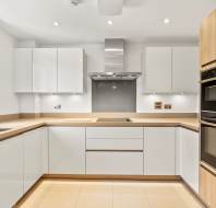 Modern kitchen with white cabinets, light wood accents, built-in double oven, electric cooktop, stainless steel hood, and a window above a single-basin sink.