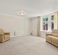 A sparsely furnished living room with beige carpet, a matching sofa and armchair, large window with curtains, and neutral-colored walls.