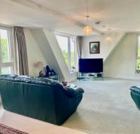 Spacious living room with large windows, two dark green sofas, a TV on a glass stand, and neutral carpeted flooring. Natural light fills the room.