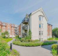 A modern apartment building with balconies, surrounded by landscaped gardens and walking paths under a clear blue sky.