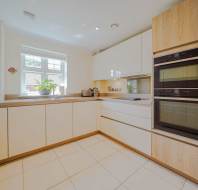 Modern kitchen with light wood and white cabinets, built-in double oven, induction cooktop, potted plant on the counter, and a window letting in natural light.