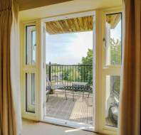 Open glass doors with beige curtains lead to a balcony with a bench and metal railing, overlooking trees and a sunlit outdoor view.