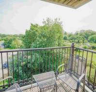 A balcony with two metal chairs and a small table overlooks a green landscape with trees, grassy areas, and parked cars on a sunny day.