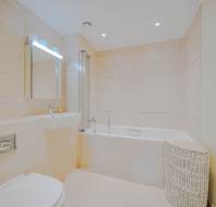 Modern bathroom with beige tiles, a built-in bathtub with glass screen, wall-mounted toilet, large mirror, soap dispenser, and a wicker laundry basket.