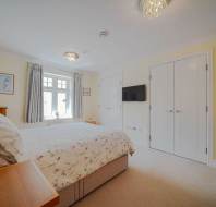 A bright bedroom with a double bed, floral bedding, a side table, TV mounted on the wall, two white closets, and a window with curtains letting in natural light.