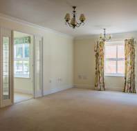 Empty living room with cream walls, beige carpet, two ceiling lights, floral curtains on windows, and double mirrored doors on the left side.
