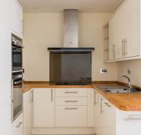 Modern kitchen with white cabinets, wood countertops, built-in oven, electric stovetop, metal range hood, and stainless steel sink against a beige wall.