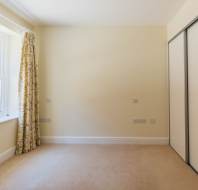 Empty room with beige carpet, cream walls, a window with floral curtains on the left, and a sliding white closet door on the right.