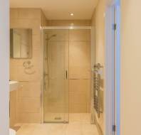 Bathroom with beige tiles, glass-enclosed shower, wall-mounted mirror, towel rail, and a white sink partially visible on the left.