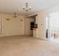 Unfurnished living room with beige carpet, cream walls, ceiling lights, and open access to a kitchen and sunroom through glass doors.