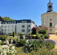 A white building with multiple windows stands beside a white clock tower, surrounded by landscaped gardens and flowering plants under a clear blue sky.
