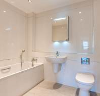 Modern bathroom with a white bathtub, wall-mounted sink, toilet, and a mirror cabinet above the sink, all set against light-colored tiled walls and floor.