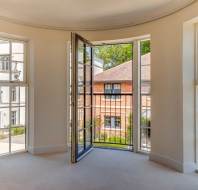 A bright, empty room with beige carpet and three tall windows, one of which is a door opening to a small balcony overlooking neighboring houses.