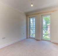 Empty room with cream-colored walls and carpet, a ceiling light, and two glass doors leading to a small balcony with views of greenery outside.