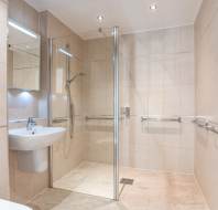 Modern bathroom with beige tiles, a walk-in glass shower, wall-mounted sink, chrome towel rail, and grab bars. Ceiling spotlights provide illumination.