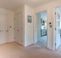 A hallway with beige carpet, white walls, three doors leading to different rooms, and a view into a kitchen and a living area.