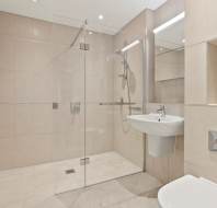 Modern bathroom with beige tiles, a glass-enclosed walk-in shower, wall-mounted sink, and built-in shelving. Toilet partially visible in the foreground.