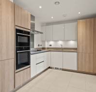 Modern kitchen with light wood and white cabinetry, built-in oven and microwave, electric cooktop, and under-cabinet lighting. Neutral tile floor and minimalistic design.