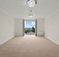 Empty, carpeted living room with cream walls, two ceiling lights, a radiator, and large glass doors leading to a balcony with a view of trees and sky in the distance.