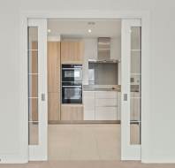 Modern kitchen with wood and white cabinets, built-in ovens, and a stainless steel hood, viewed through open sliding glass doors.