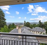 View from an open window showing rooftops of neighboring houses, trees, and a distant landscape under a blue sky with scattered clouds.