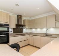 Modern kitchen with beige cabinets, built-in oven, electric cooktop, stainless steel vent hood, sink, and ample countertop space under bright overhead lighting.