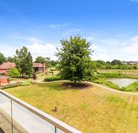 View from a balcony overlooking a large lawn with a central tree, a pond, footpaths, landscaped gardens, and several houses under a blue sky with scattered clouds.