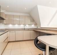 Modern kitchen with beige cabinets, built-in oven, induction cooktop, under-cabinet lighting, and a curved breakfast bar with a black stool. Light-colored tile flooring and neutral countertops.