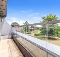 A modern balcony with glass railing overlooks a landscaped area with trees, grass, and distant houses under a clear blue sky.