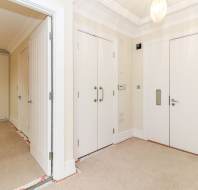 A clean hallway with beige carpet, white walls, and several closed white doors, including a main entrance door with a security handle.