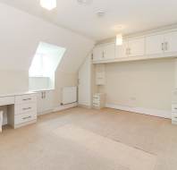 A spacious, unfurnished room with built-in white cabinets, desk, a small stool, beige carpet, and a window allowing natural light.