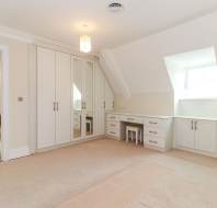 Spacious bedroom with built-in white wardrobes, a dressing table, beige carpet, sloped ceiling, and a window letting in natural light.
