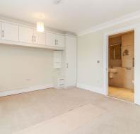 Unfurnished bedroom with built-in white cabinets, beige carpet, overhead light fixture, and open door leading to a tiled bathroom.