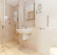 Modern accessible bathroom with beige tiles, a wall-mounted sink, walk-in shower with glass door, grab bars, mirror cabinet, and a toilet partially visible on the right.