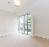 Empty carpeted room with cream walls, ceiling light, and glass doors leading to a balcony with trees visible outside.