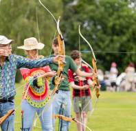 A group of people standing in a line outdoors, aiming bows and arrows, with trees and people in the background.