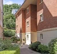 A paved walkway runs between two modern apartment buildings with red brick and cream facades, surrounded by neatly trimmed bushes and greenery.