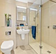 Modern bathroom with beige tiles, wall-mounted toilet and sink, glass shower enclosure, mirror cabinet, and blue towels on a rail. Flowers and toiletries are arranged on shelves.