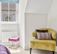 A yellow velvet armchair with two cushions sits next to a window; a pink stool is nearby, and part of a bed and dresser are visible in a bright, modern room.